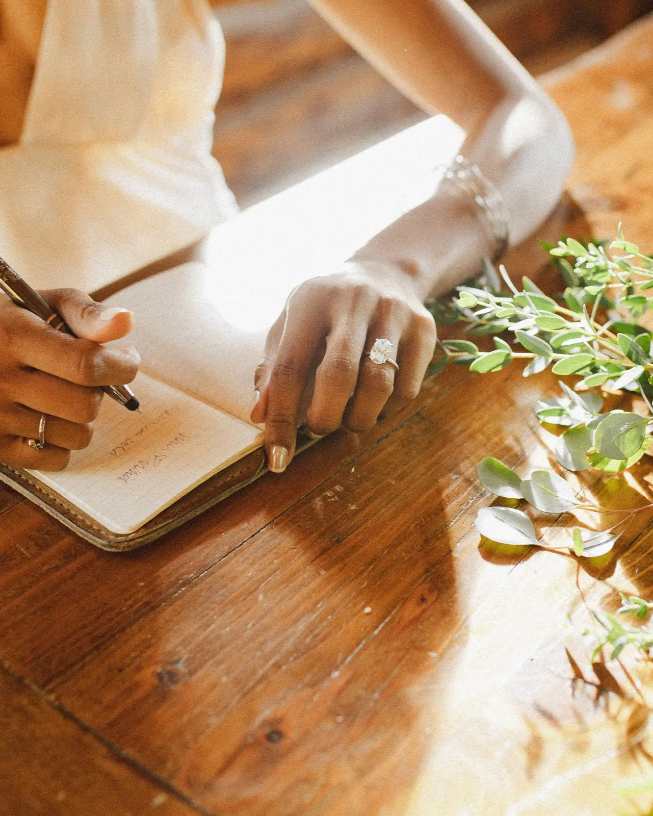 Hands writing wedding vows at a wooden table in soft window light