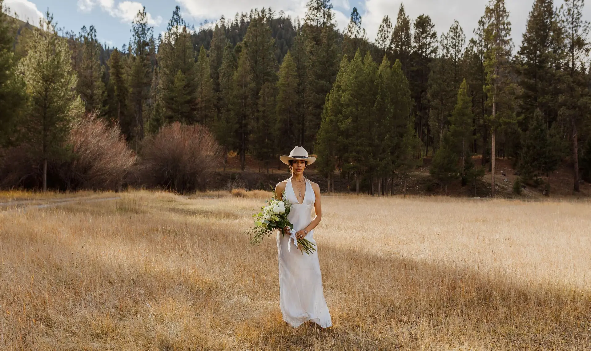 A bride in a cowboy hat holding her bouquet in the golden meadow, with the pine forest behind