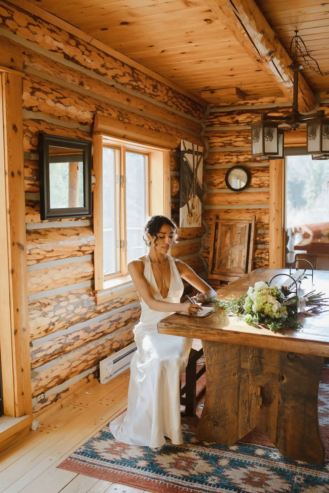 A bride in a silk gown writing at a wooden table inside a log cabin
