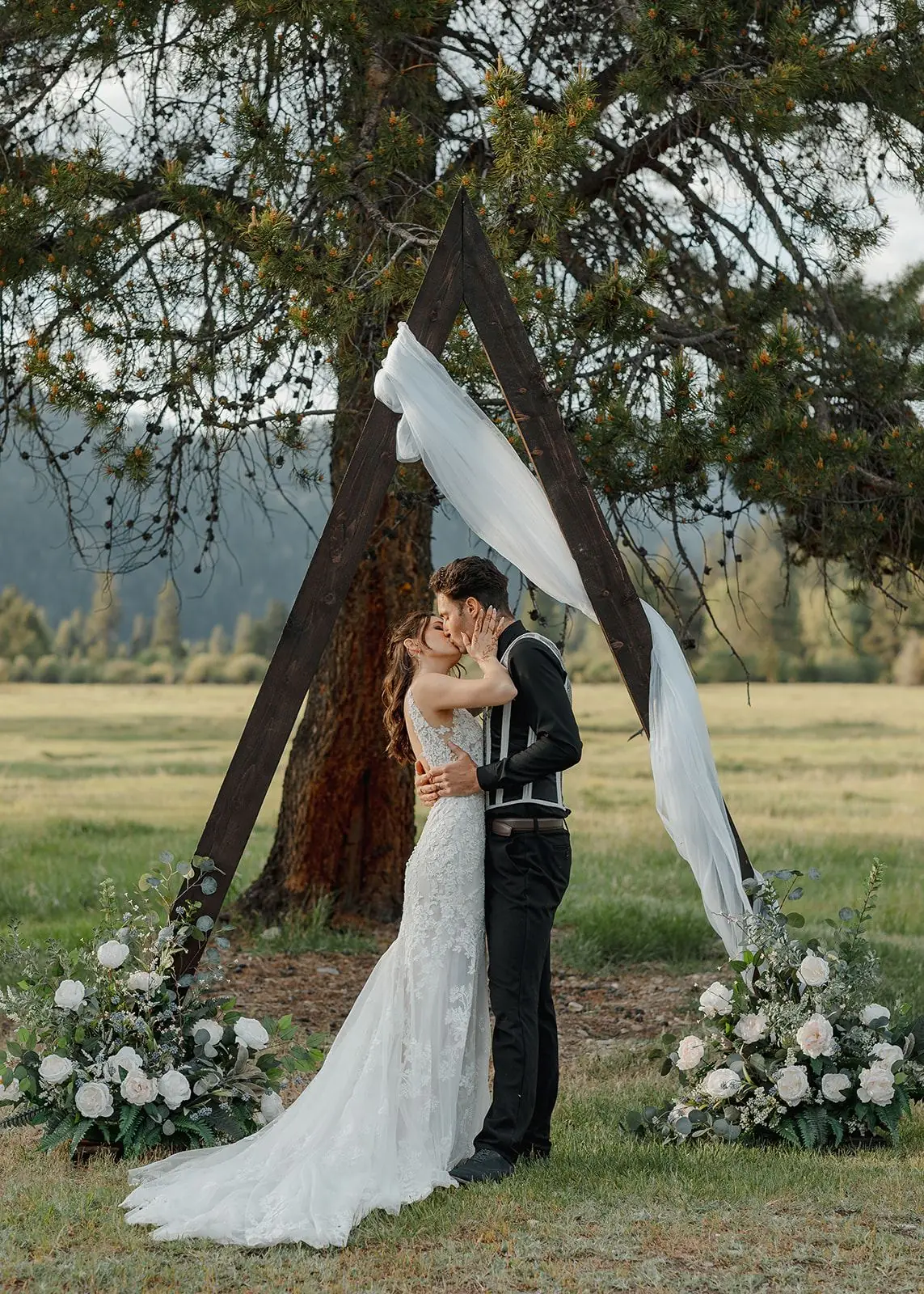 A bride and groom kiss under a florals-draped wooden arch beside a lone pine at Alta Ranch