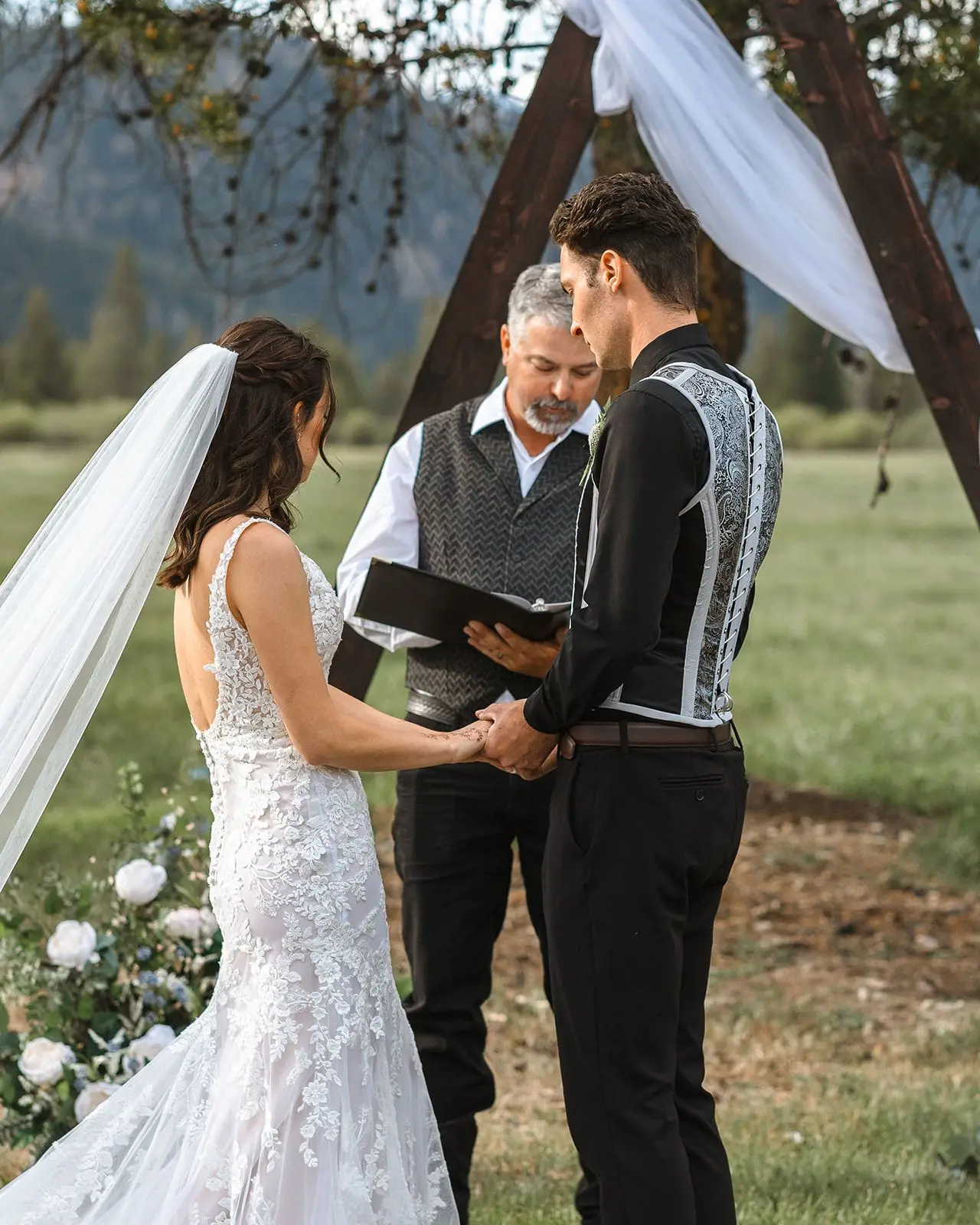 A bride and groom holding hands during their vows at a wooden arch in the meadow