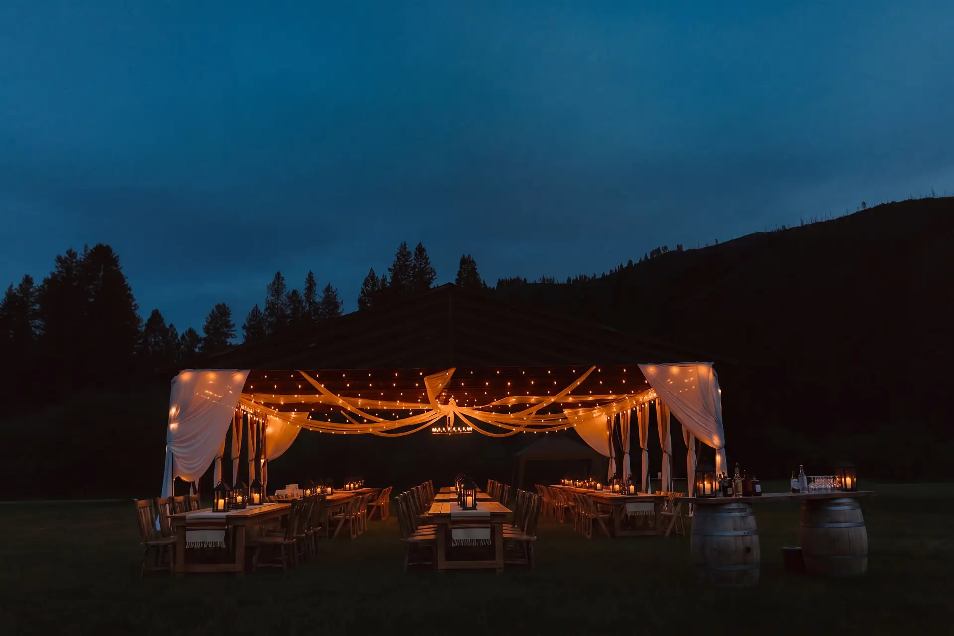 The historic hangar lit up at night, string lights and white drapes glowing over long tables against the dark hills