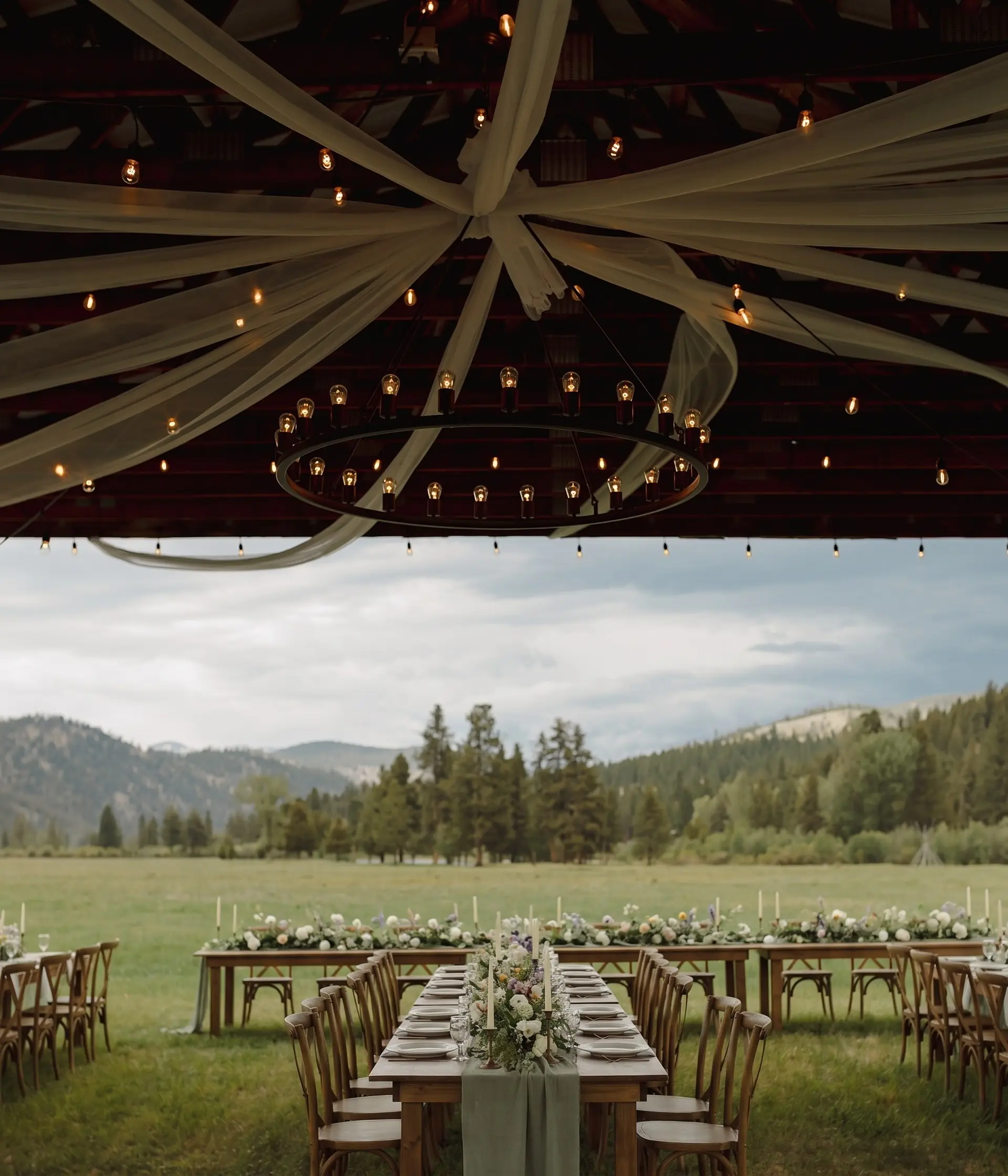 Styling idea: a long dinner table set with florals under the hangar roof, looking out over the meadow to the pines